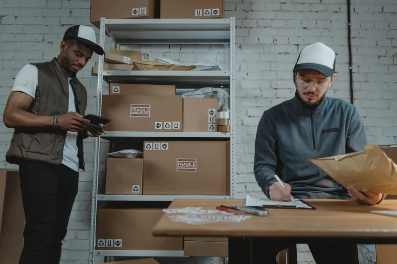 Two warehouse workers checking inventory boxes on shelving, one using a tablet and the other writing on a clipboard