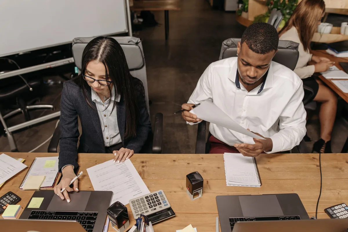 Two office employees at a desk reviewing printed procedure documents alongside open laptops and notebooks